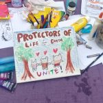 A child's painting that reads "Protectors of Life and Earth Unite!" with animals with hearts over their heads, between two trees