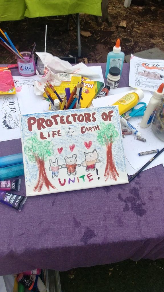 A child's painting that reads "Protectors of Life and Earth Unite!" with animals with hearts over their heads, between two trees