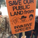 Person holding sign that reads "Save our public land from invasive species" with pictures of Bundy family